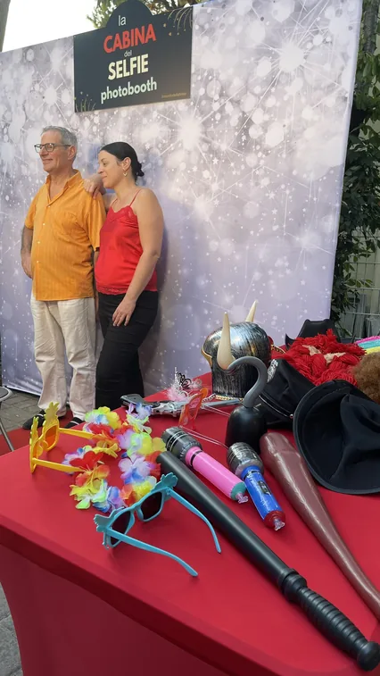 Un uomo in camicia arancione e una donna in canotta rossa in posa davanti al fondale argentato con l'insegna La Cabina del Selfie; in primo piano il tavolo rosso con occhiali colorati, collane di fiori, microfono gonfiabile, elmo e cappelli