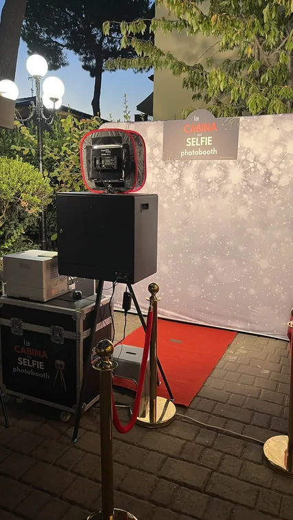 La colonna del photobooth vista da vicino: la fotocamera dentro il diffusore rosso, la stampante appoggiata sul baule con il logo La Cabina del Selfie e, alle spalle, il fondale argentato con il tappeto rosso